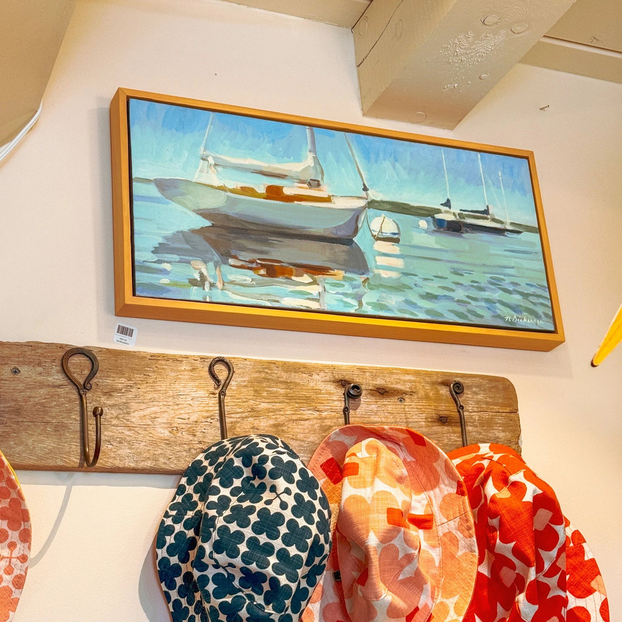 Framed painting of sailboats on a wall above a wooden hat rack with colorful hats.